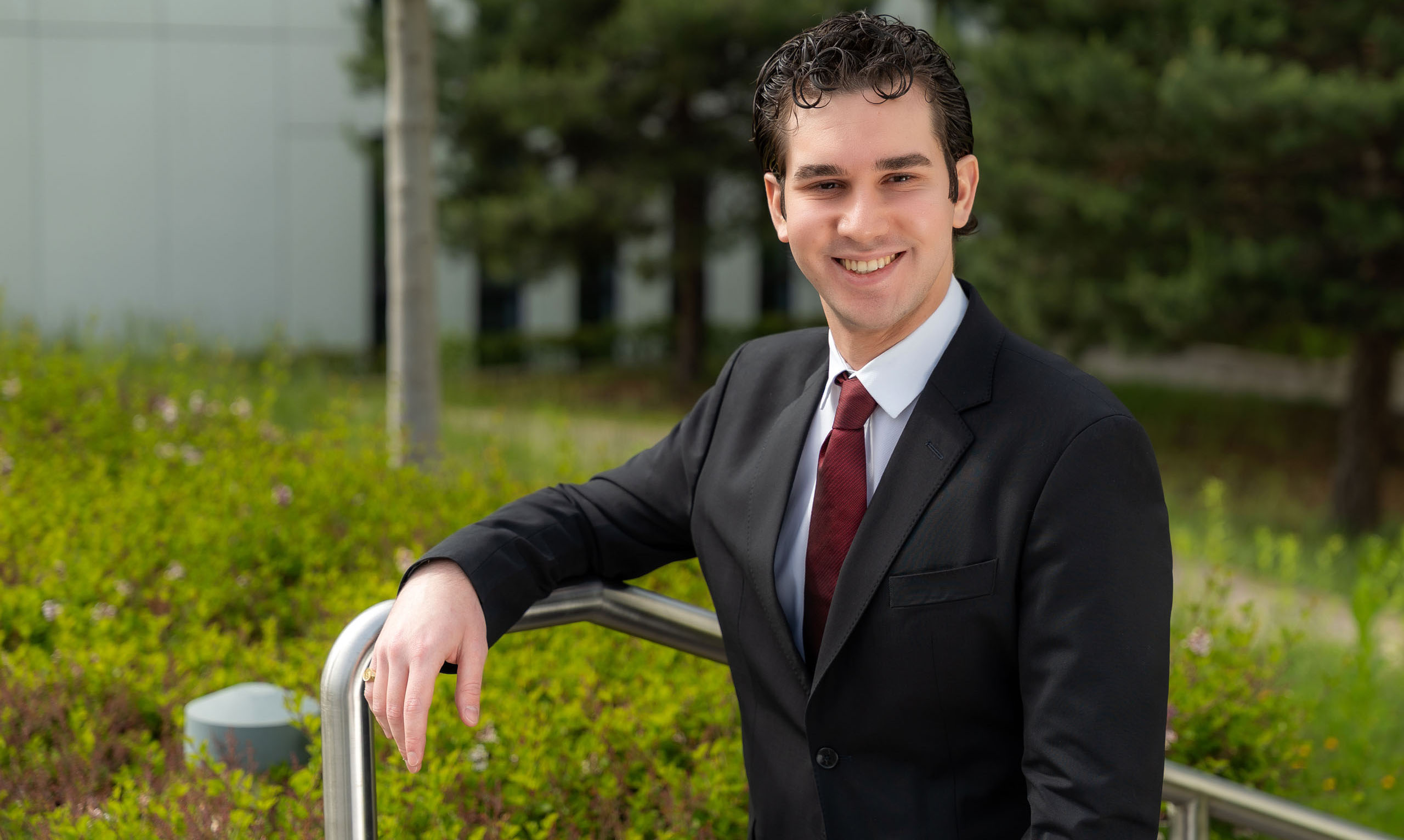 A man wearing a black jacket, white shirt, and burgundy tie is standing outside, leaning against a metal railing. In the background, green bushes, trees, and part of a building are visible.