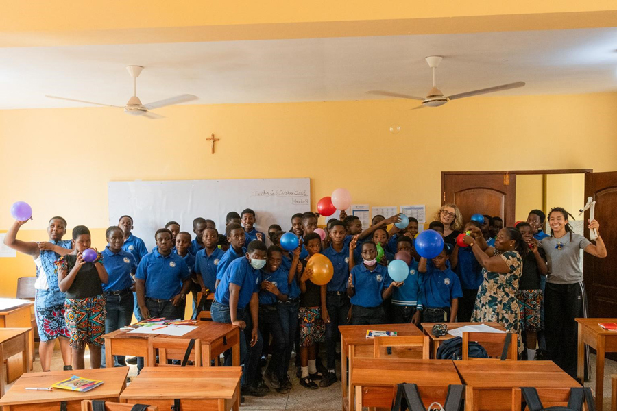 Około 30 osób w sali lekcyjnej pozuje do zdjęcia. Część z nich trzyma kolorowe balony: niebieskie, fioletowe, pomarańczowe i różowe. Wszyscy się uśmiechają. Większość ubrana jest w niebieskie koszulki. Przed nimi stoją drewniane ławki, na których leżą przybory lekcyjne. Za nimi widać tablicę lekcyjną.