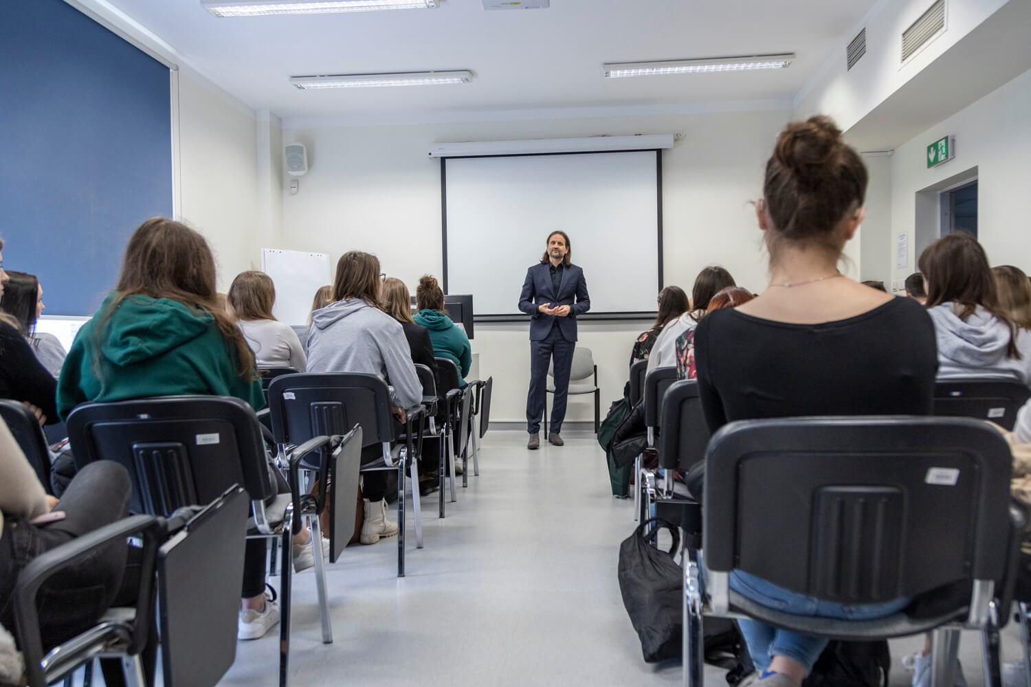 A group of people in a lecture theatre. They are sitting on chairs. In front of them stands a middle-aged man. He is dressed in a suit.