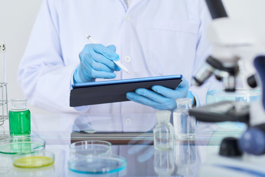 naukowiec w laboratorium, widać tylko biały fartuch i niebieskie rękawiczki, zapisuje coś w tablecie, na stole szklane naczynia laboratoryjne