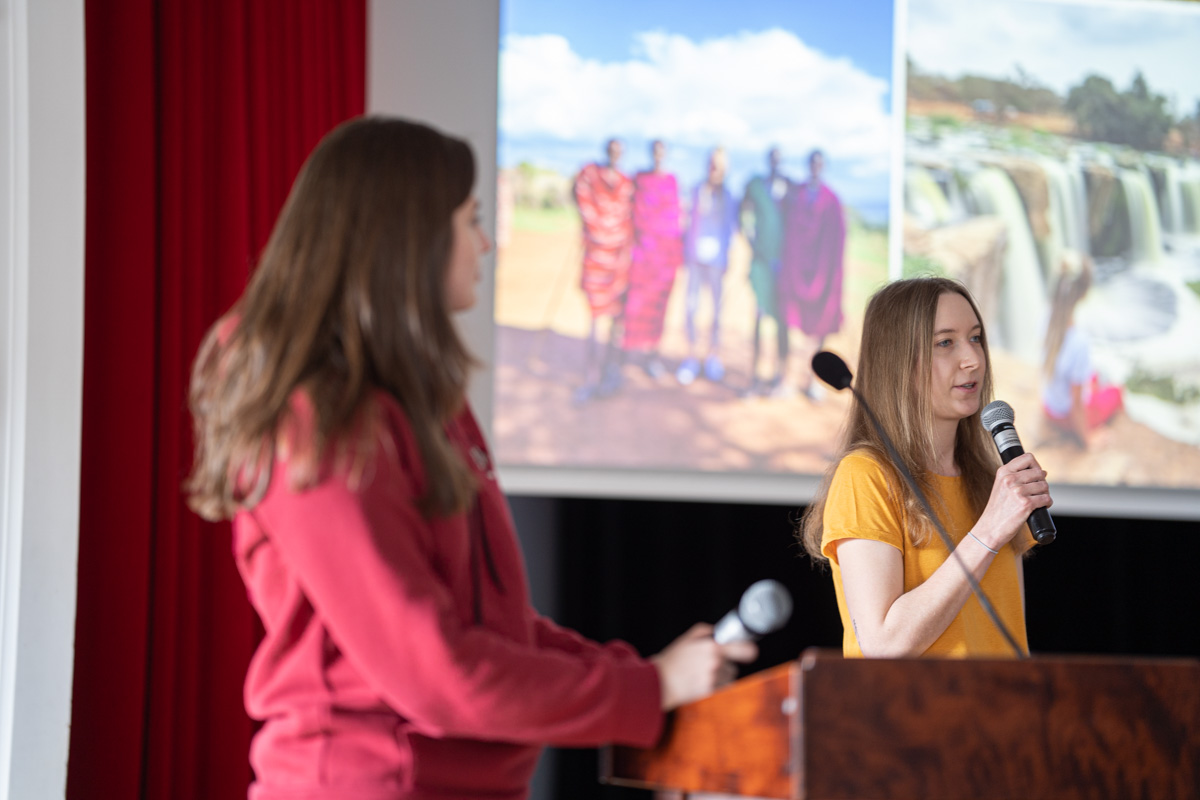 Dwie młode dziewczyny przy mównicy. Pierwsza w czerwonej bluzie, druga w żółtej bluzce z krótkim rękawem. Ta druga mówi do mikrofonu. W tle na ekranie zdjęcia z Afryki.