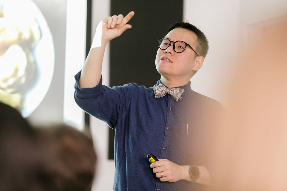 A man of Asian descent wearing glasses and a bow tie with his hand raised