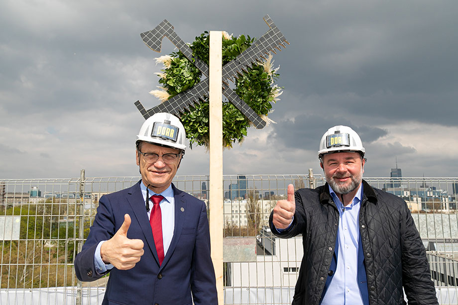 Two men wearing helmets on the roof of a building with their arms outstretched and thumbs up, behind them thye toppng out