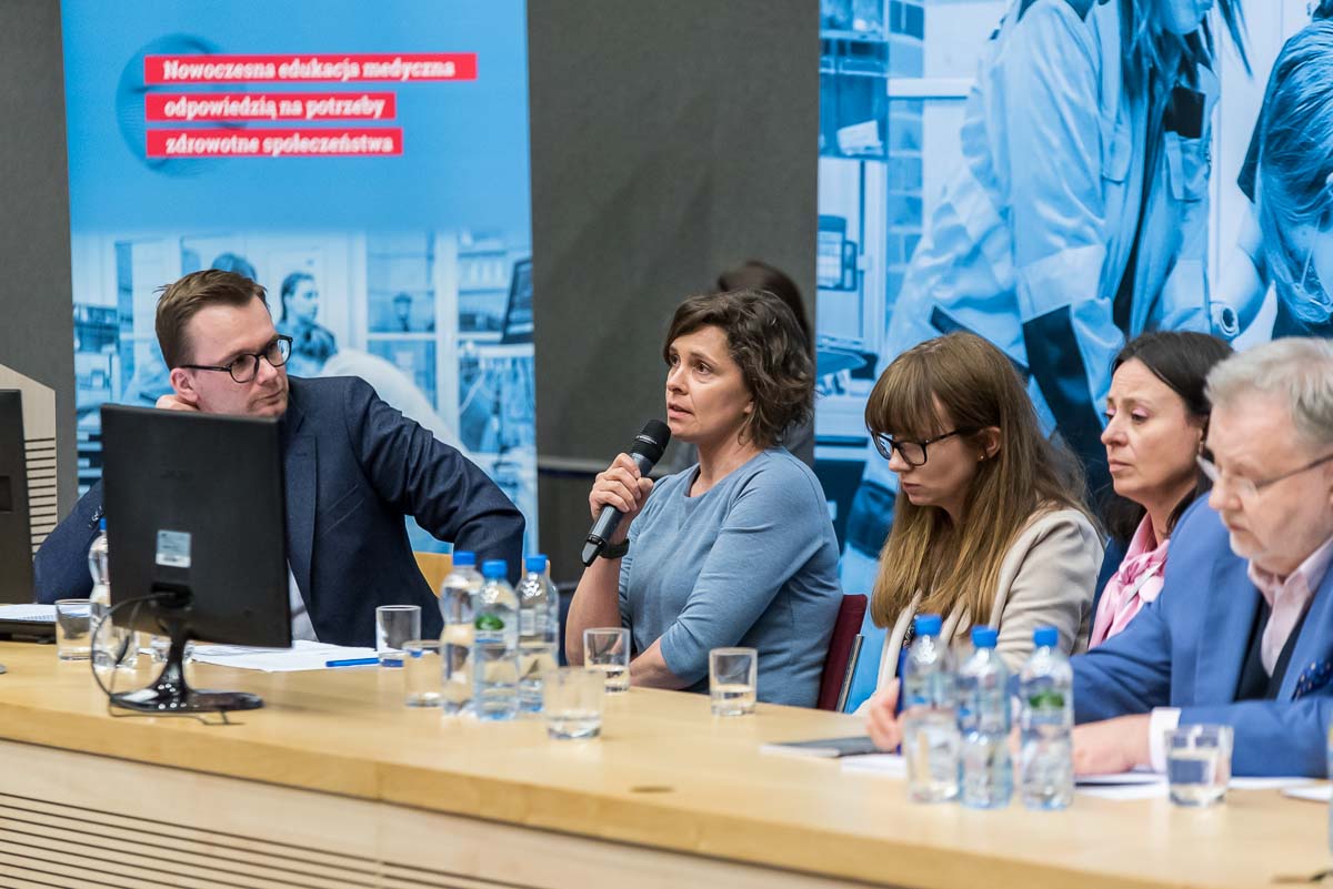 Five people are seated at a table. The second from the right - a woman in a blue dress - speaks into a microphone.