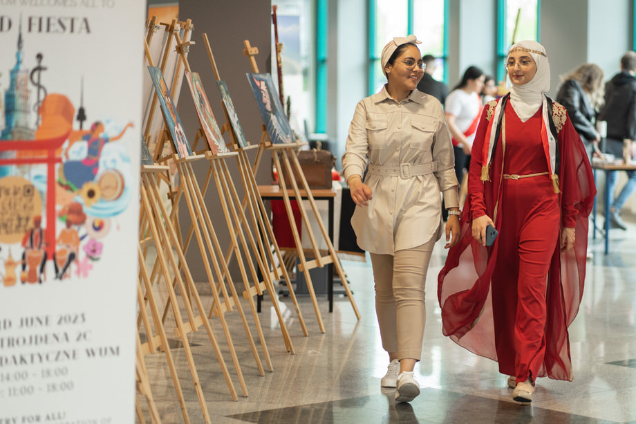 Two female students are walking along a corridor where an exhibition is taking place. On the right you can see easels on which paintings have been set up. The girl on the left in traditional Arab dress and a white hijab on her head. The girl on the right dressed in safari style.