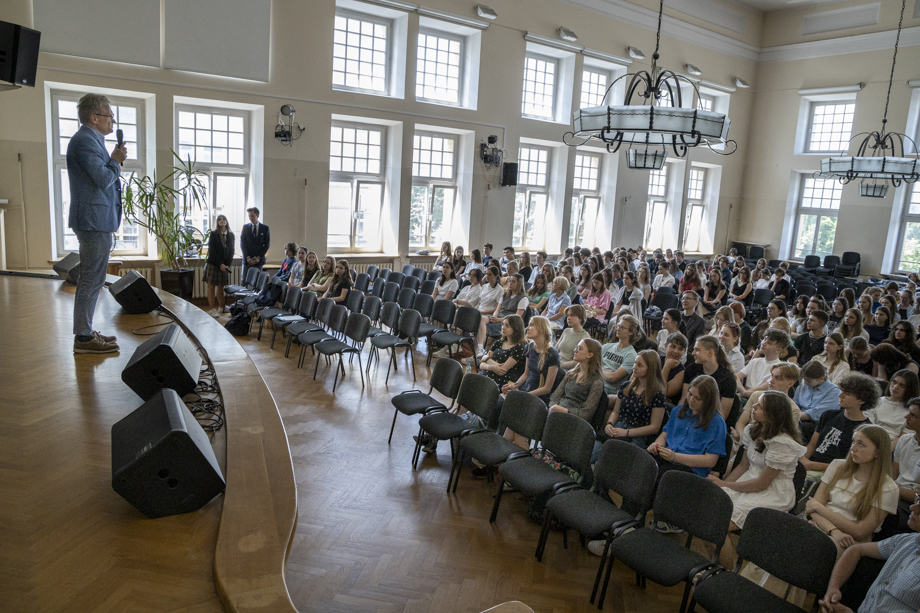 Lecture hall. At the lectern, a man in a blue suit speaks into a microphone. In the audience a lot of young people.