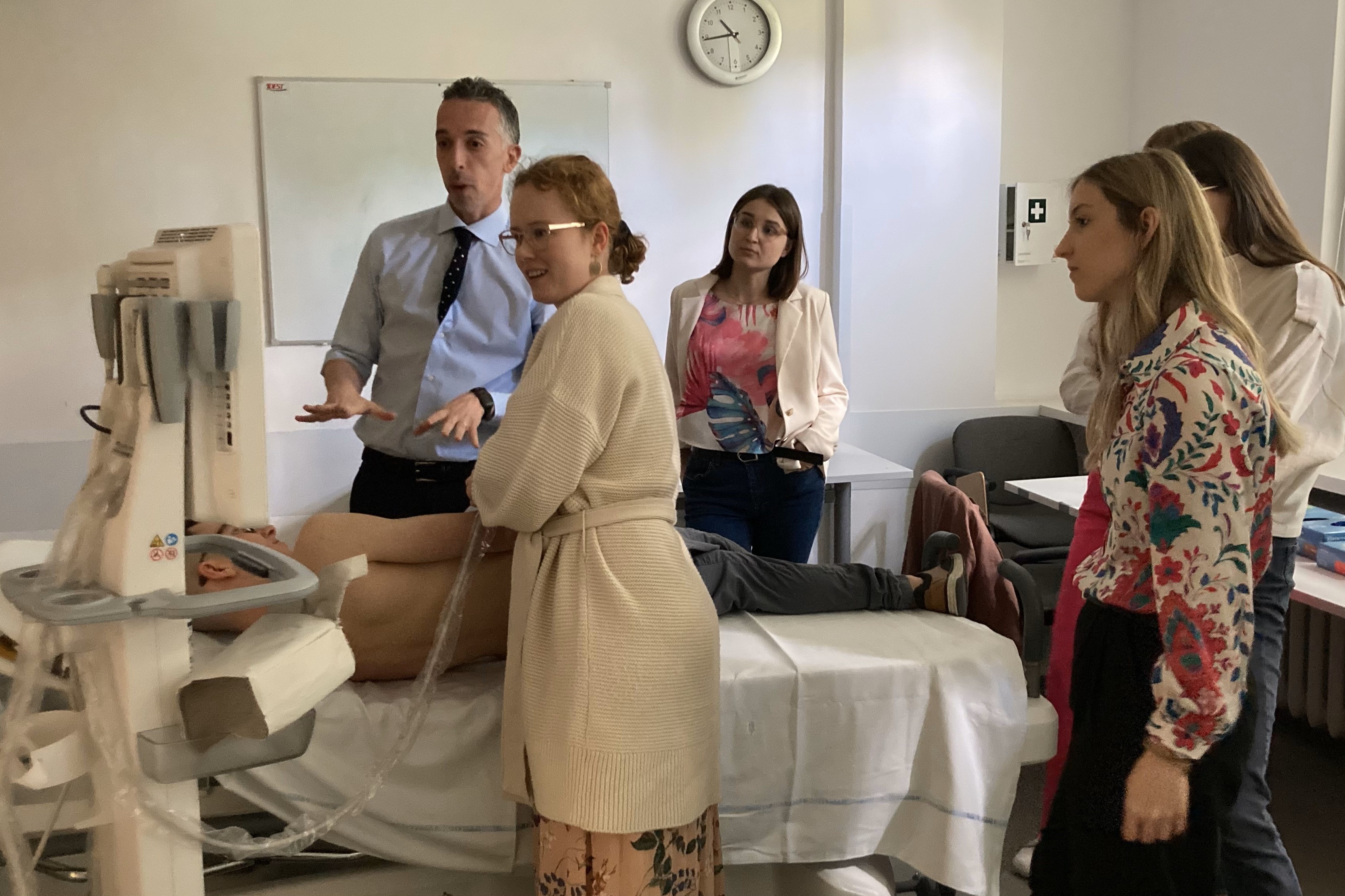 The lecturer - a tall slim man in a blue shirt and six students at the ultrasound camera. Next to the apparatus is a hospital bed on which one of the students lies undressed from the waist up. A female student is performing an ultrasound on him.