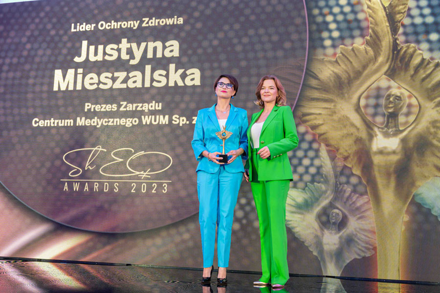 Dwie stojące na scenie kobiety. Jedna ubrana w niebieski garnitur, druga w zielony. Kobieta po lewej trzyma statuetkę. Za nimi ekran mlultimedialny.
