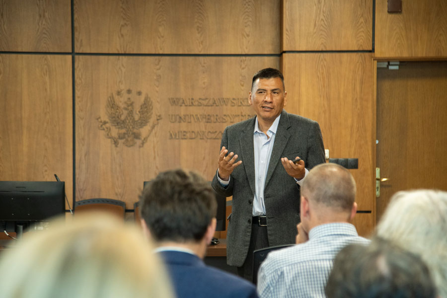 A man dressed in a suit stands in the middle of the room. He has his hands raised and is speaking to the audience. The audience sits in front of him, with their backs to the camera.