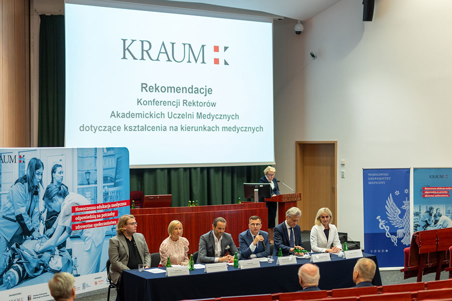 A group of people sitting behind a table: two women and four men. Next to them rollups. Behind them a screen with the KRAUM logo. In the back, a woman is standing on a podium behind a lectern, speaking.