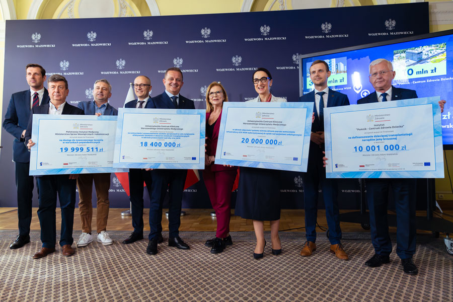 A group of people, men and women. They are standing. They are smiling for a photograph. In their hands they hold large cardboard cheques with the amount and name of the grant in blue. 