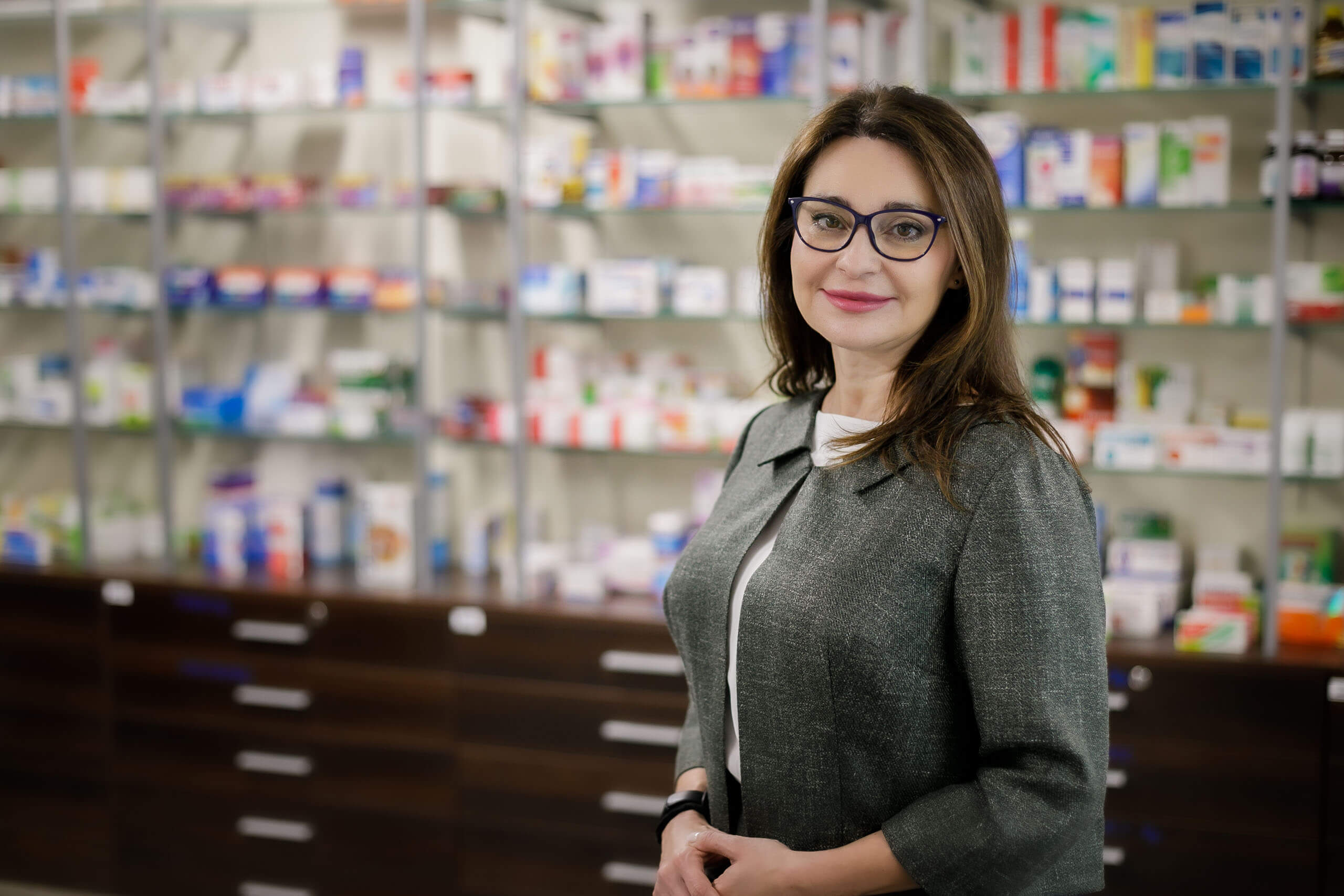 Na pierwszym planie kobieta w okularach, w tle apteczne półki z lekami.