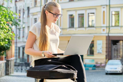 A young girl with glasses sits on a wall in the city with a yellow tenement in the background.