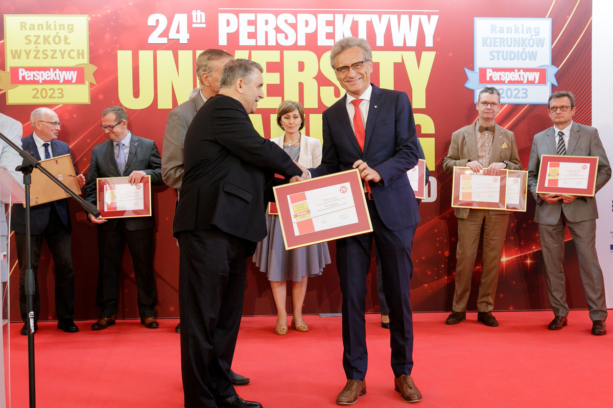 Two three men in the foreground. On the left, a short man in a suit hands the man in a suit, standing on the right, a diploma. Behind them, a group of people.