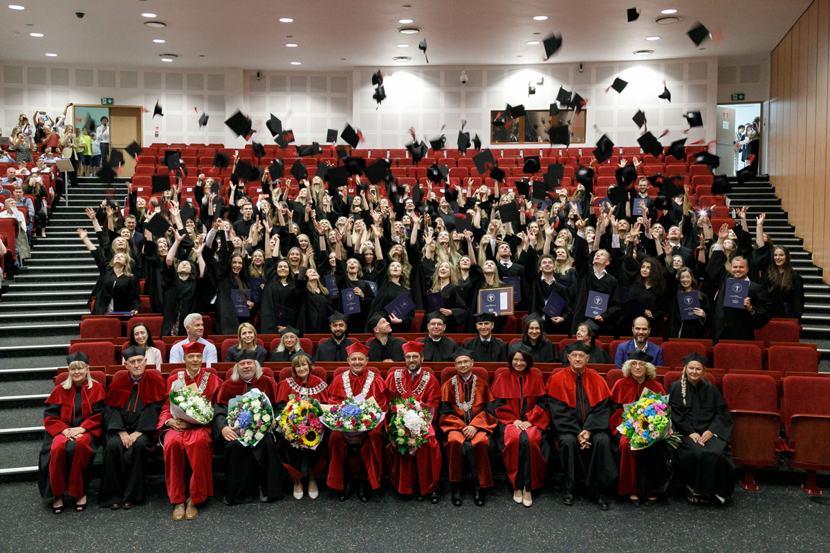 Lecture hall. In the front rows sit teachers in academic togas. In the back stand graduates in student togas - they throw their birettas up in the air.