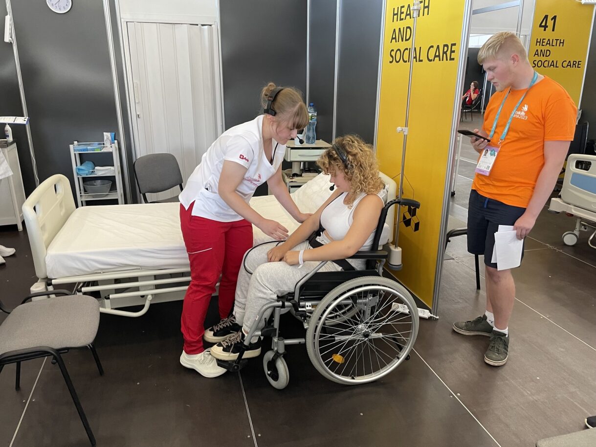 A young girl nurse leans over a woman sitting in a wheelchair. In the back is a man in an orange T-shirt.