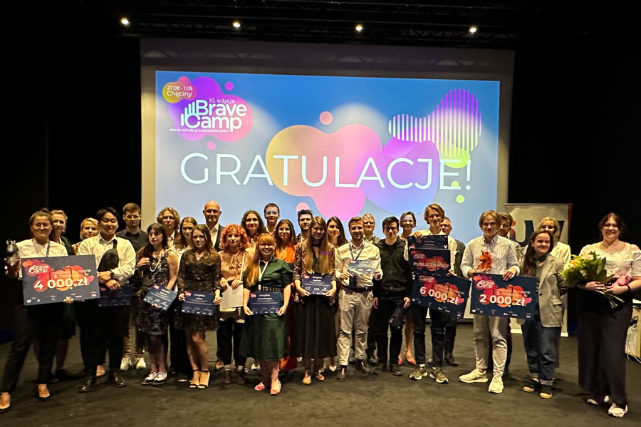 A group of young people standing with diplomas, flowers. Behind them a screen displaying the name Bravecamp