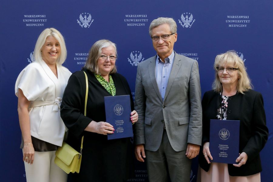 NN Four people are standing in front of a banner with the logos of the Warsaw Medical University. Three women and a man. All of them are wearing elegant clothes. Two women are holding navy blue briefcases with the WUM logo