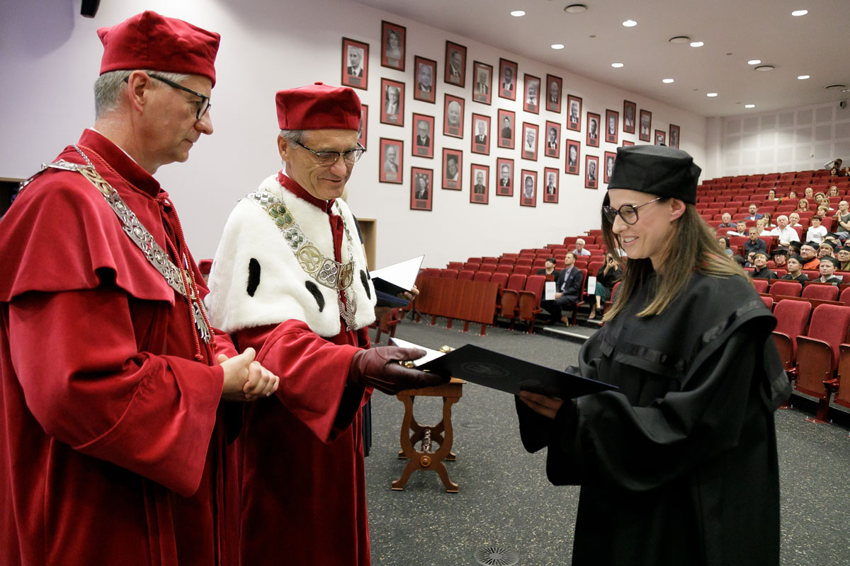 Dwóch mężczyzn i kobieta w togach na auli. Mężczyzna w czerwonej todze wręcza dyplom kobiecie w czarnej todze.