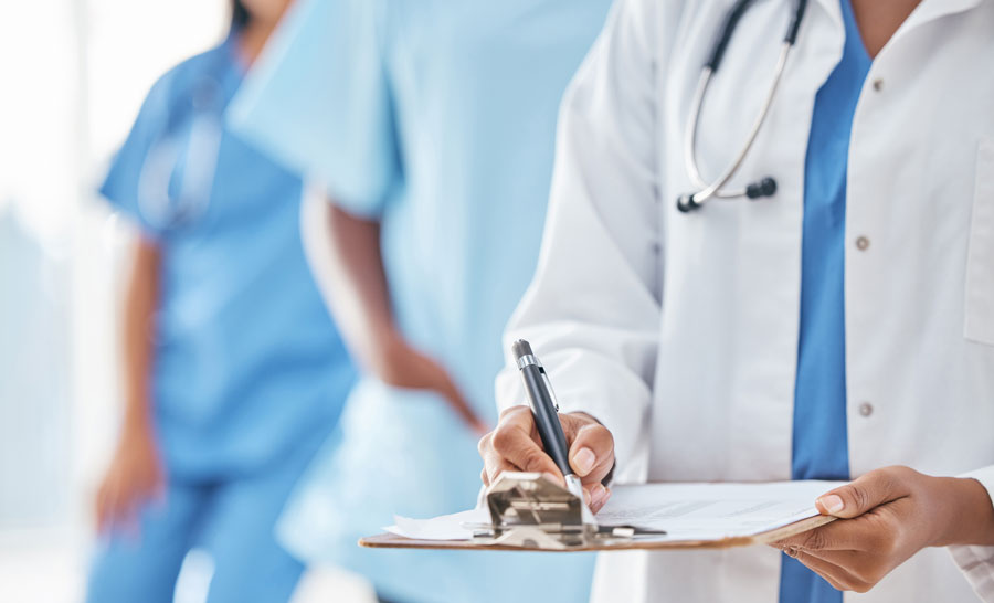 Three people wearing medical gowns: white, blue and blue, the person in the foreground is taking notes and has a stethoscope hanging around his neck