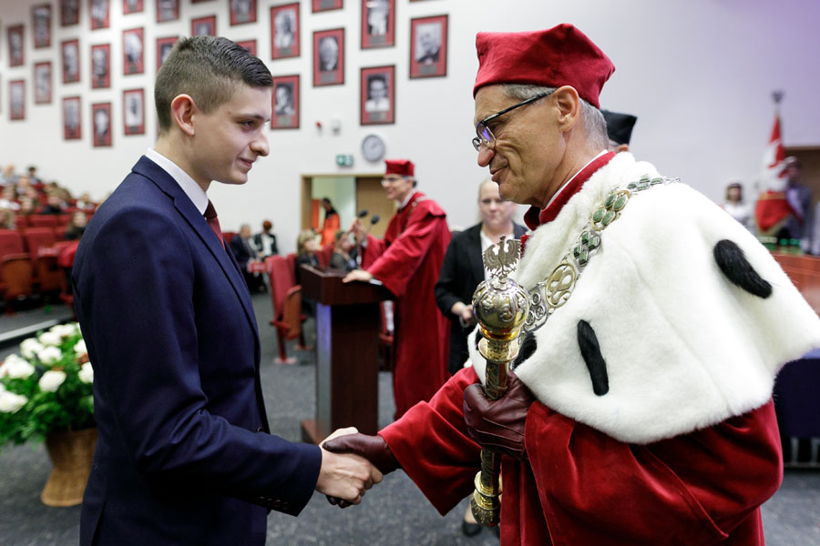 On the left a young man in a suit, on the right a middle-aged man in a rector's toga - red and white touches the young man's shoulder with a scepter.