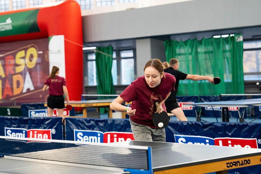 Dziewczyna ubrana na sportowo pochyla się nad stołem do pingponga, w jednej ręce trzyma paletkę, za nią w tle - młodzi sportowcy