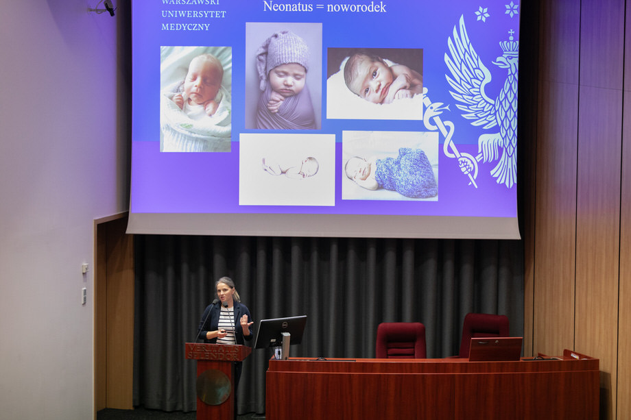 Kobieta stojąca za mównicą, za nią ekran multimedialny ze slajdem w kolorze niebieskim i ze zdjęciami noworodków