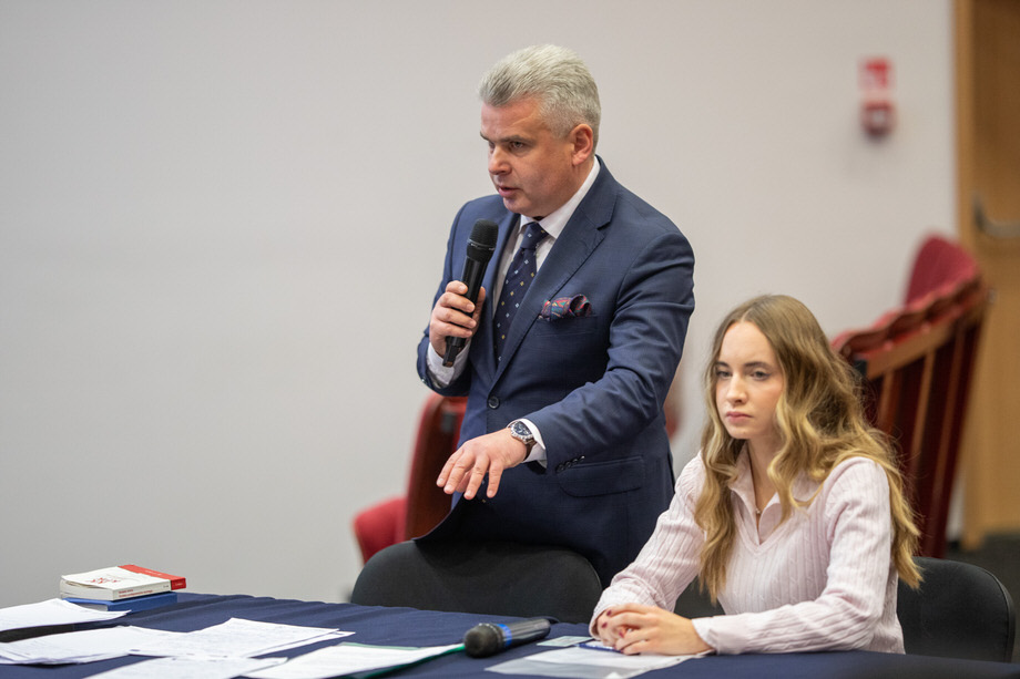 Przy stole siedzi młoda dziewczyna. Obok niej starszy mężczyzna w garniturze stoi i mówi do mikrofonu.