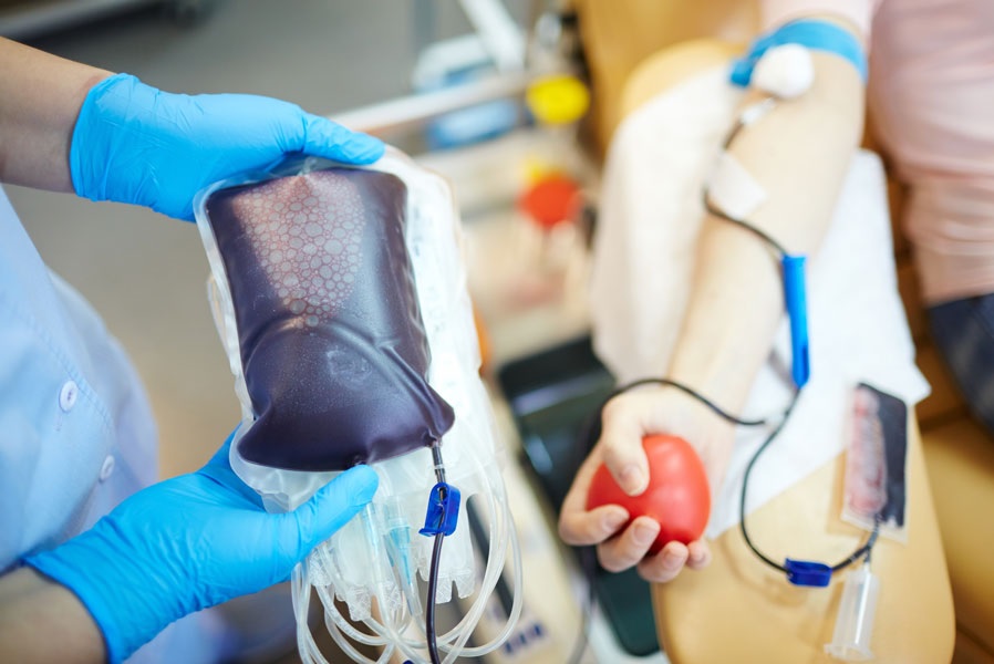 Pacjent w czasie badania hematologicznego, ma podłączoną krew, widać jego rękę ściskającą czerwoną piłkę relaksacyjną oraz dłonie pielęgniarki w niebieskich rękawiczkach, które trzymają worek z krwią.