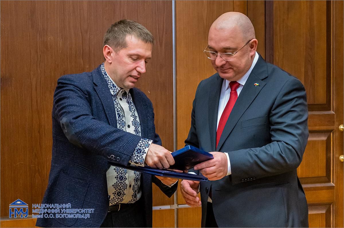 Dwóch mężczyzn w garniturach, jeden przekazuje drugiemu dyplom i medal