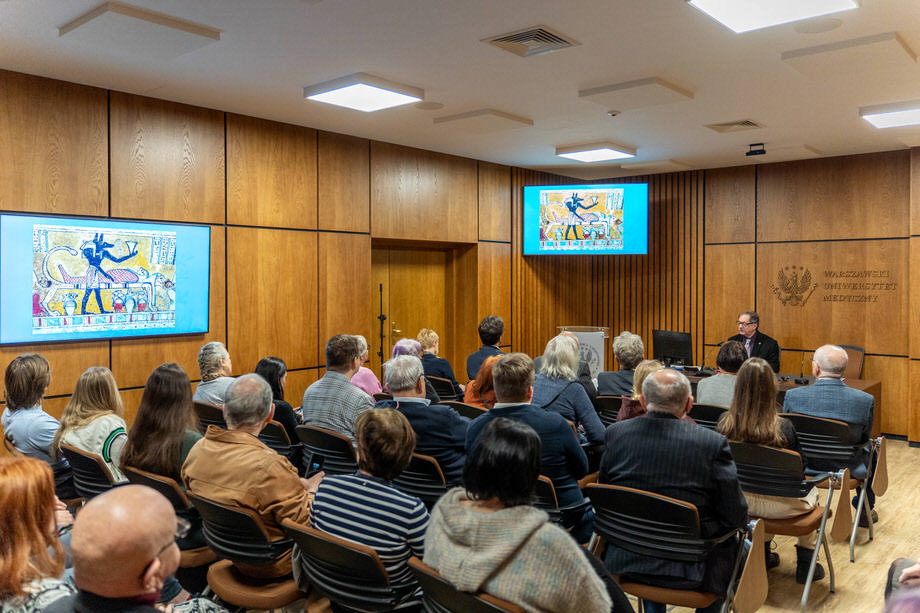 Lecture hall. Two screens showing Egyptian engravings. The audience visible from the back. At the lectern table - the speaker.