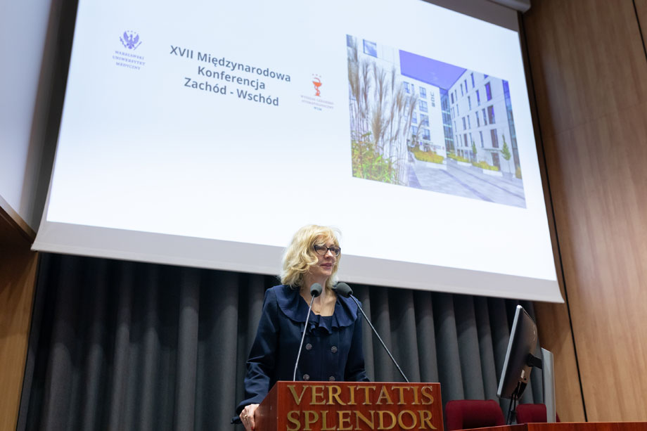 Elegancko ubrana kobieta, podsekretarz stanu w Ministerstwie Zdrowia, stoi za mównicą i przemawia. Za nią ekran multimedialny, na nim zdjęcie białego budynku i tytuł konferencji