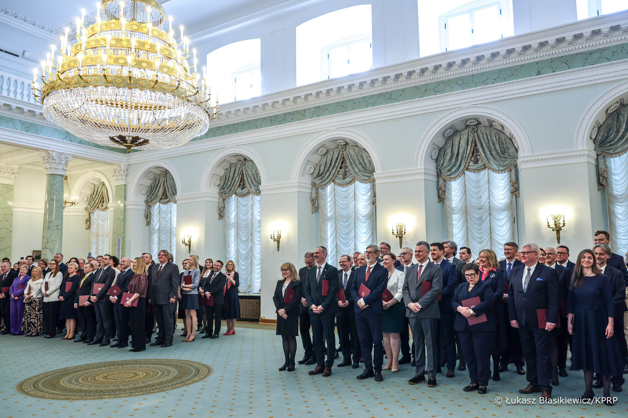 Grupa osób elegancko ubranych. Stoją w dużej okazałej sali.