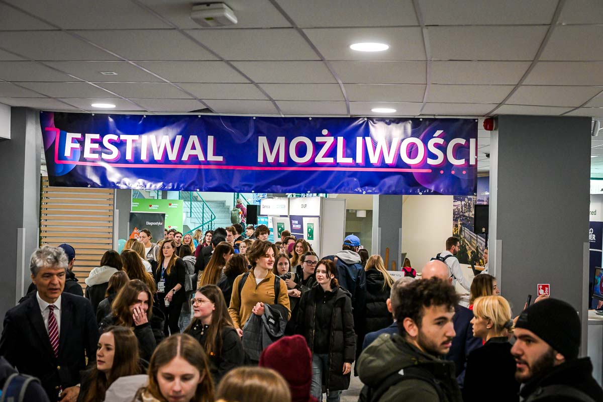 Tłum ludzi, uczestników wydarzenia Festiwalu Możliwości. Znajdują się na korytarzu, na którym są też różne stoiska wystawiennicze. Nad nimi wisi fioletowy baner z napisem Festiwal Możliwości.