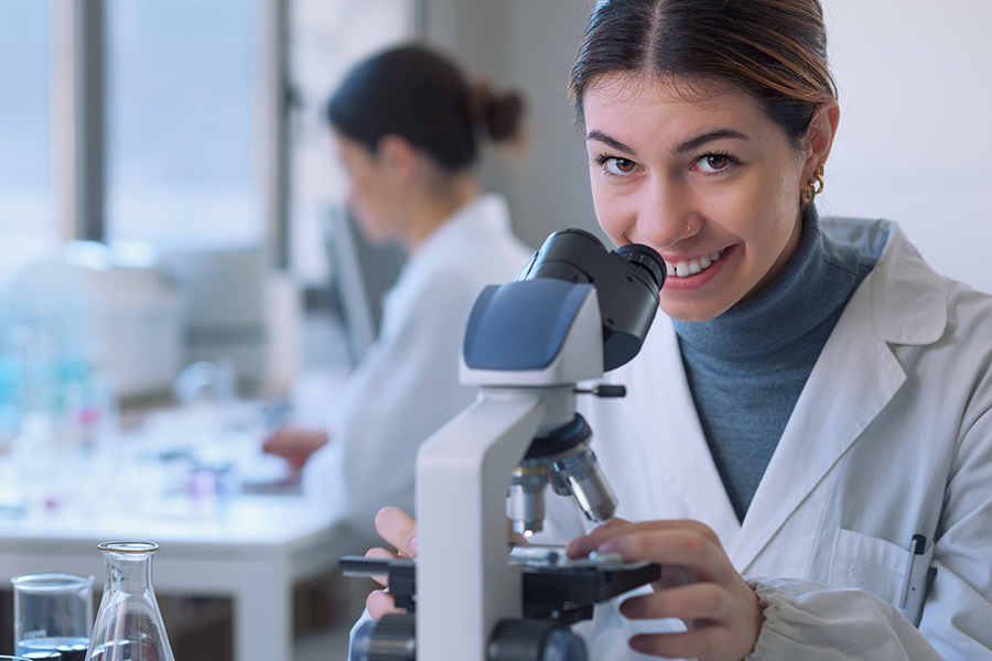Na obrazie widzimy dwie kobiety w laboratorium. Kobieta na pierwszym planie używa mikroskopu. Uśmiecha się do zdjęcia. Kobieta w tle pracuje przy biurku z różnym sprzętem laboratoryjnym. Obie osoby mają na sobie białe fartuchy laboratoryjne. Na stole widoczne są również szklane naczynia laboratoryjne.