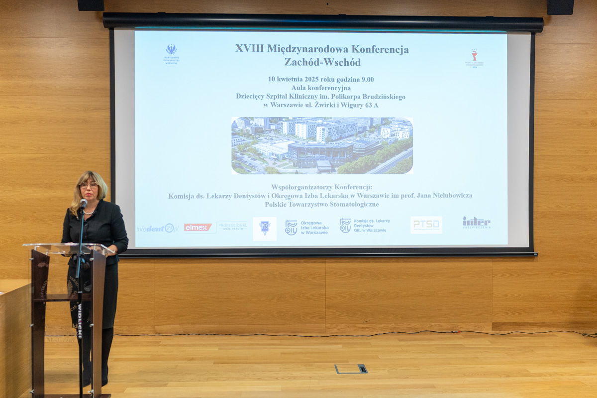 An elegantly dressed woman - Prof. Agnieszka Mielczarek, dean of WLS. She stands at the lectern and welcomes the conference guests. Behind her is a large screen with the name of the conference, a photo of the Banach campus and the organizers and co-organizers listed.