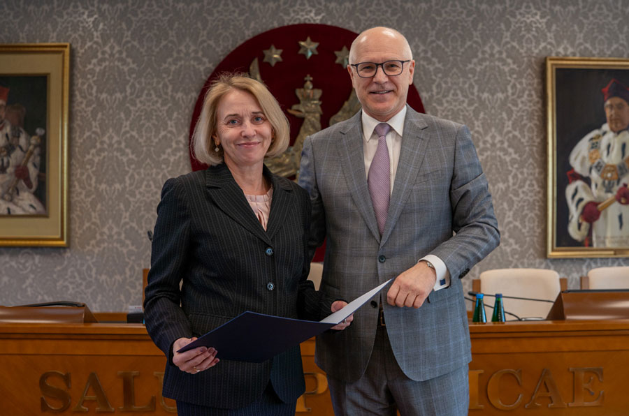 Kobieta i mężczyzna stojący obok siebie. Kobieta po lewej ubrana jest w ciemny garnitur i trzyma przed sobą niebieską teczkę. Mężczyzna po prawej ma na sobie szary garnitur w kratę oraz jasnofioletowy krawat. Uśmiechają się do zdjęcia. Za nimi widać drewniany stół. W tle znajduje się czerwone godło WUM z orłem i gwiazdami. 