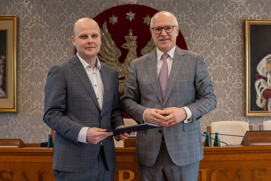 Dwóch mężczyzn stojących obok siebie. Obaj mają na sobie szare garnitury i białe koszule; mężczyzna po lewej nie ma krawata, natomiast mężczyzna po prawej ma. Uśmiechają się do zdjęcia. Za nimi widać drewniany stół. Mężczyzna po lewej trzyma teczkę, natomiast mężczyzna po prawej ma splecione dłonie. Za nimi jest ściana z tapetą z motywem roślinnym i czerwonym godłem WUM.