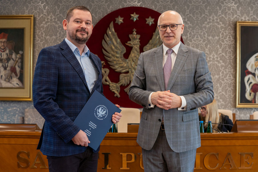 Dwóch stojących obok siebie mężczyzn. Mężczyzna po lewej ubrany jest w niebieski garnitur i jasnoniebieską koszulę. Trzyma w rękach niebieską teczkę. Mężczyzna po prawej ma na sobie szary garnitur, białą koszulę i jasnofioletowy krawat. Ma splecione dłonie. Obaj uśmiechają się do zdjęcia.