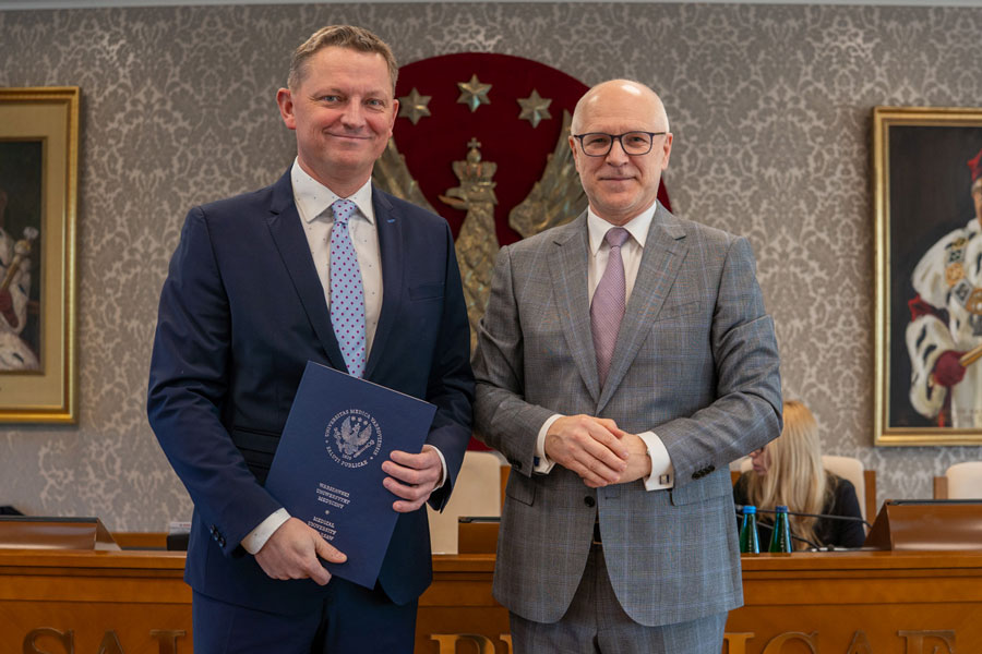 Dwóch stojących obok siebie mężczyzn. Mężczyzna po lewej ubrany jest w niebieski garnitur i jasnoniebieską koszulę oraz krawat. Trzyma w rękach niebieską teczkę. Mężczyzna po prawej ma na sobie szary garnitur, białą koszulę i jasnofioletowy krawat. Ma splecione dłonie. Obaj uśmiechają się do zdjęcia.