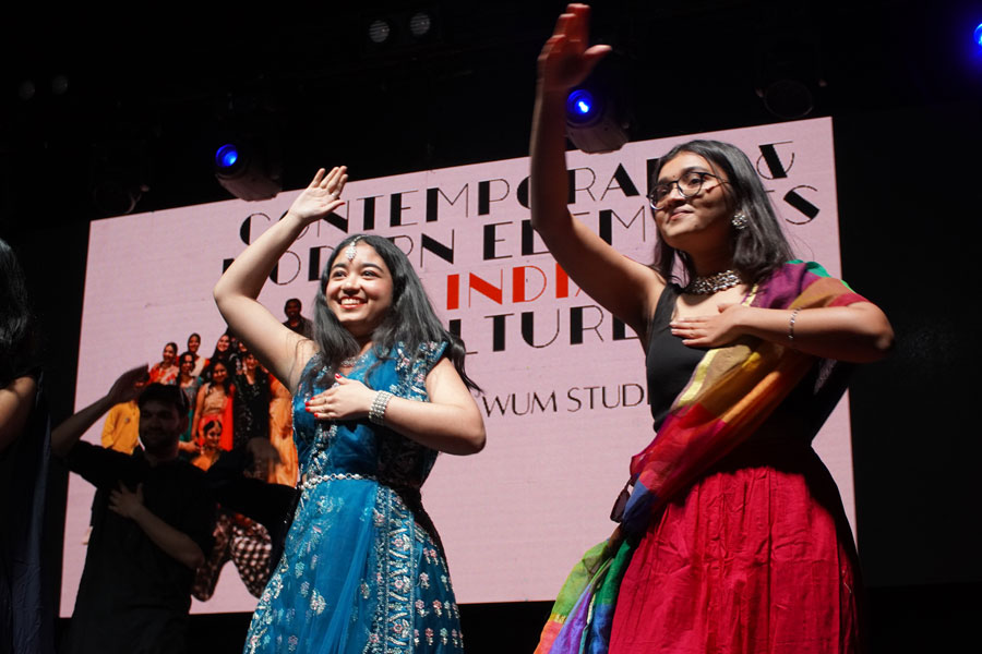 A dance scene featuring a group of people. In the foreground are two women performing a dance in traditional Indian costumes. The woman on the left is wearing a long blue dress decorated with embroidered patterns. The woman on the right is wearing a red, flared skirt and a black blouse with a colorful shawl draped over it. Both dancers have their arms raised in dynamic poses. A projection screen with subtitles can be seen in the background. The entire stage is intensely lit. Translated with DeepL.com (free