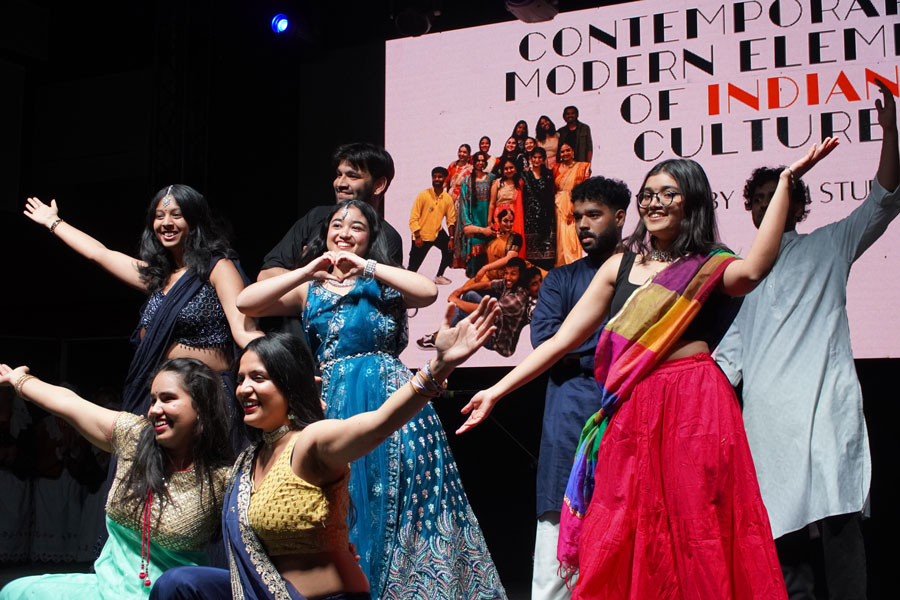 A group of young people - WUM students - standing on stage. They are wearing colorful traditional costumes. They make various gestures with their hands and pose for a photo. In the background is a large screen with the caption “Contemporary modern elements of Indian culture”.