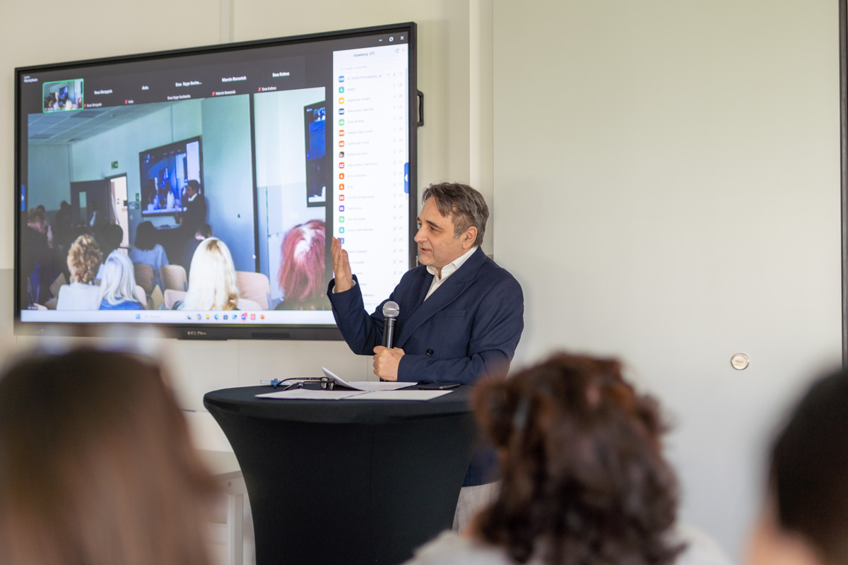 A man - Dr. Piotr Dziechciarz - in a suit stands behind a black lectern, holding a microphone and gesturing with his hand. Behind him is a large screen displaying a video conference with many participants visible on the screen. The room is set up for a presentation or lecture, with several people seated and facing the speaker.