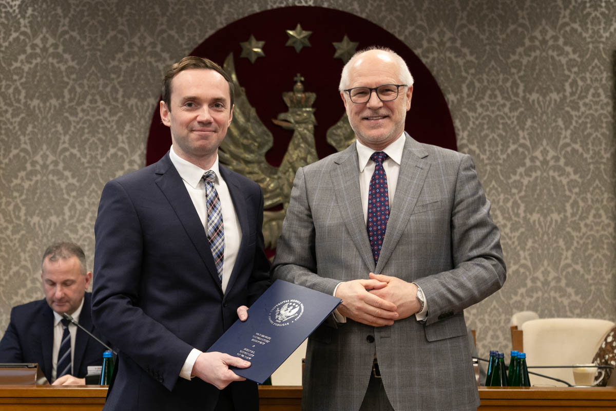 W przestronnej sali stoją obok siebie dwaj mężczyźni. Uśmiechają się do zdjęcia. Mężczyzna po lewej ma ciemny garnitur i krawat i trzyma niebieską teczkę z godłem i nazwą WUM. Mężczyzna po lewej ma szary garnitur i ciemny krawat. W tle widoczny jest duży herb z orłem i gwiazdami na czerwonym tle oraz osoby siedzące przy stole. 