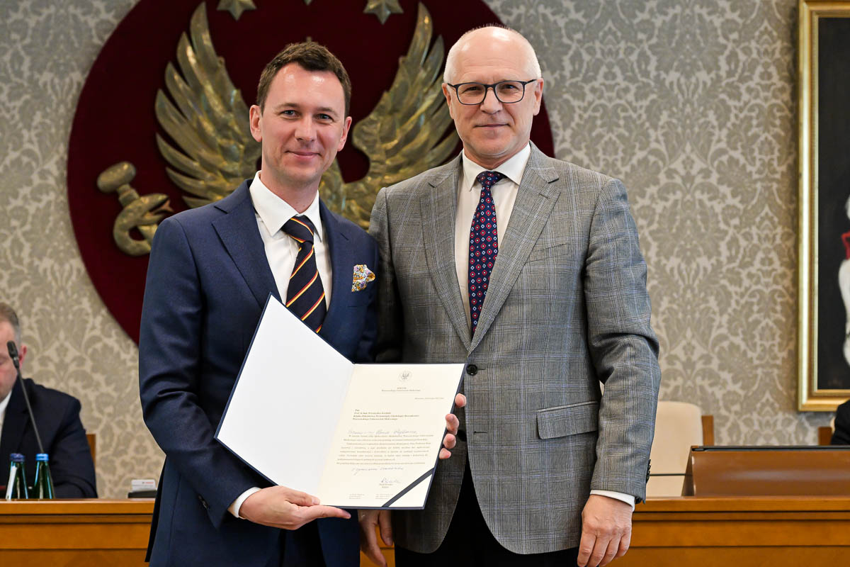 Dwóch mężczyzn w formalnych strojach, stojących obok siebie i uśmiechających się do zdjęcia. Mężczyzna po lewej stronie ubrany jest w granatowy garnitur, białą koszulę i krawat w paski. Trzyma otwartą teczkę z dokumentem. Mężczyzna po prawej stronie ma na sobie szary garnitur w kratę, białą koszulę i krawat w czerwono-niebieskie kropki. W tle znajduje się duże godło WUM z białym orłem na czerwonym tle. Obok herbu widoczny jest fragment obrazu w złotej ramie.