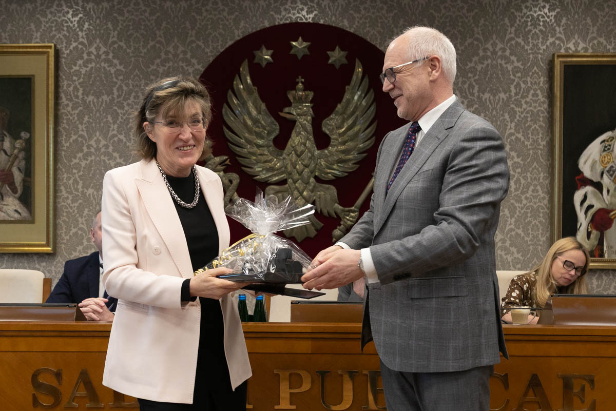 W przestronnej sali stoi obok siebie kobieta i mężczyzna. Kobieta stoi po lewej. Ma jasną marynarkę i czarne spodnie. Trzyma w rękach prezent, opakowany w przezroczystą folię z kokardą. Mężczyzna po prawej nosi szary garnitur. Obie osoby uśmiechają się. W tle widoczny jest czerwony herb z białym orłem oraz fragmenty dwóch obrazów na ścianie. Za mężczyzną i kobietą widać inne osoby, które siedzą.