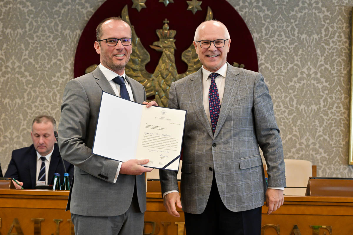 Dwóch mężczyzn w garniturach, stojących obok siebie i uśmiechających się do zdjęcia. Mężczyzna po lewej stronie trzyma otwartą teczkę z dokumentem. Mężczyzna po prawej stronie ma na sobie szary garnitur w kratę, białą koszulę i krawat w czerwono-niebieskie kropki.  W tle znajduje się duże godło WUM z białym orłem na czerwonym tle. Obok herbu widoczny jest fragment obrazu w złotej ramie.