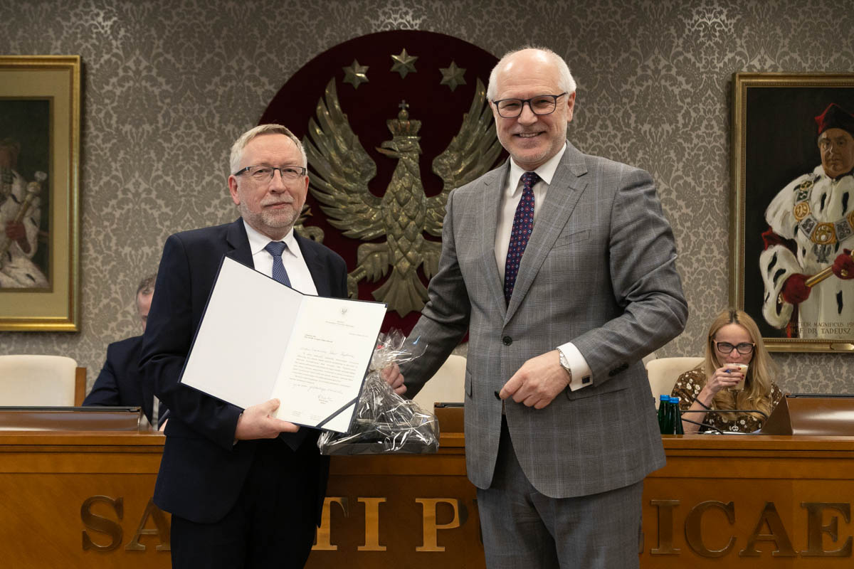 W przestronnej sali stoi obok siebie dwóch mężczyzn. Mężczyzna po lewej ma ciemny garnitur i koszulę w paski, a po prawej – szary garnitur i krawat w kropki. Mężczyzna po lewej stronie ubrany jest w ciemny garnitur i ciemny krawat, a mężczyzna po prawej w szary garnitur i krawat w kropki. Mężczyzna po lewej trzyma w rękach otwartą teczkę z dokumentem. W tle, na czerwonym tle, widoczny jest duży herb z białym orłem, a obok niego znajduje się obraz przedstawiający postać jednego z byłych rektorów uczelni.