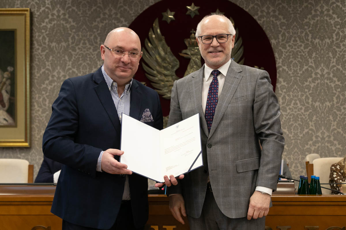 W przestronnej sali stoi obok siebie dwóch mężczyzn. Mężczyzna po lewej ma ciemny garnitur i koszulę w paski, a po prawej – szary garnitur i krawat w kropki. Mężczyzna po lewej stronie ubrany jest w ciemny garnitur i ciemny krawat, a mężczyzna po prawej w szary garnitur i krawat w kropki. Mężczyzna po lewej trzyma w rękach otwartą teczkę z dokumentem. W tle, na czerwonym tle, widoczny jest duży herb z białym orłem, a obok niego znajduje się obraz przedstawiający postać jednego z byłych rektorów uczelni.