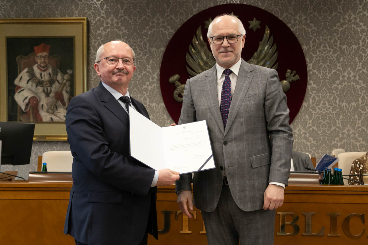 W przestronnej sali stoi obok siebie dwóch mężczyzn. Mężczyzna po lewej stronie ubrany jest w ciemny garnitur i ciemny krawat, a mężczyzna po prawej w szary garnitur i krawat w kropki. Mężczyzna po lewej trzyma w rękach otwartą teczkę z dokumentem. W tle, na czerwonym tle, widoczny jest duży herb z białym orłem, a obok niego znajduje się obraz przedstawiający postać jednego z byłych rektorów uczelni.